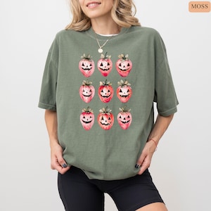 May include: Moss green t-shirt featuring a repeating pattern of strawberries with jack-o'-lantern faces. The strawberries are red and pink with green stems. The shirt is a loose fit and has short sleeves.