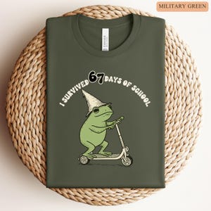 May include: Military green t-shirt with a whimsical graphic of a green frog wearing a wizard hat and riding a scooter. The text reads "I survived 67 days of school."