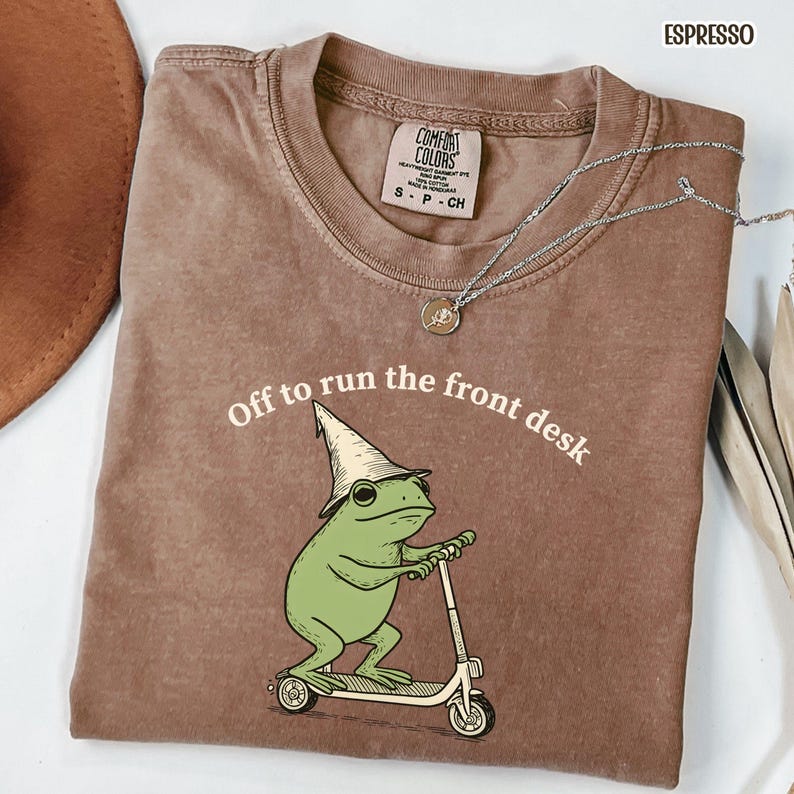 K&ouml;nnte beinhalten: Espressofarbenes T-Shirt mit dem Text "Off to run the front desk". Die Grafik zeigt einen gr&uuml;nen Frosch mit einem Zaubererhut, der einen Roller f&auml;hrt. Eine silberne Halskette mit Anh&auml;nger ist &uuml;ber dem Shirt drapiert.
