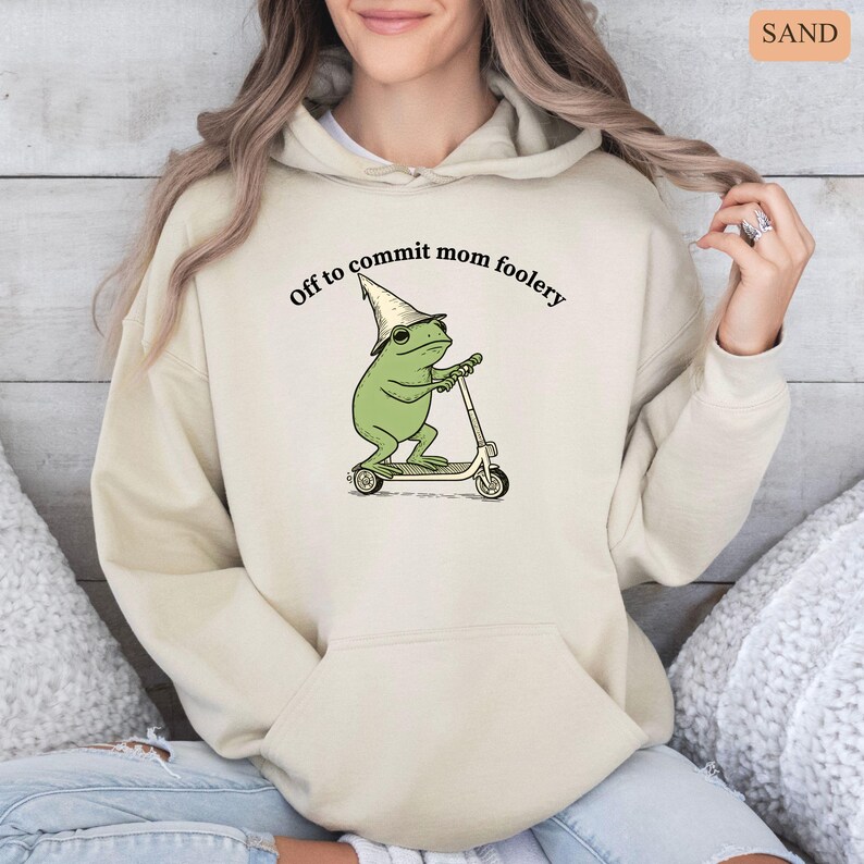 May include: Sand-colored hoodie with a front graphic of a green frog wearing a wizard hat and sunglasses, riding a scooter. The text "Off to commit mom foolery" is above the frog. The hoodie has a drawstring hood and a front pocket.