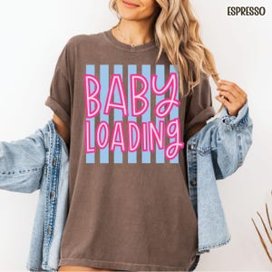 May include: Espresso-colored t-shirt with the text "BABY LOADING" in pink lettering over a light blue striped background. The shirt has short sleeves and a relaxed fit. A denim jacket is worn over the shirt.