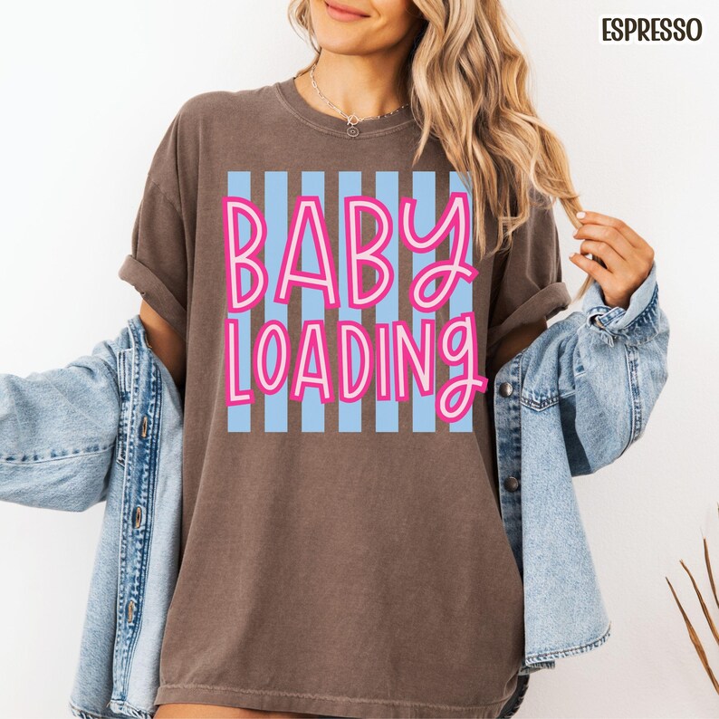 May include: Espresso-colored t-shirt with the text "BABY LOADING" in pink lettering over a light blue striped background. The shirt has short sleeves and a relaxed fit. A denim jacket is worn over the shirt.