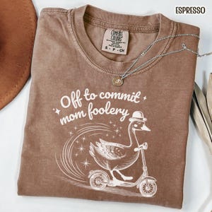 May include: Espresso-colored t-shirt with the text "Off to commit mom foolery" and a graphic of a goose riding a scooter. The shirt has a crew neck and short sleeves. A silver necklace with a pendant is on the shirt.