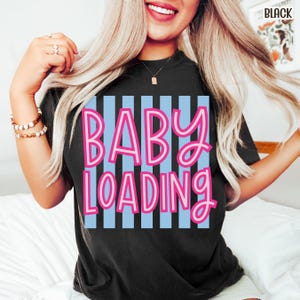 May include: Black t-shirt with the text "BABY LOADING" in pink letters over a light blue striped background. The shirt is a crew neck style.