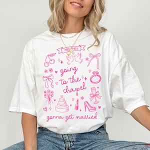May include: White t-shirt with a pink graphic design featuring wedding-themed illustrations and text. The design includes cherries, doves, bows, a ring, a cake, lipstick, a high heel, and the phrases "going to the chapel" and "gonna get married".