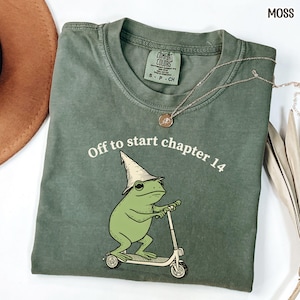 May include: Moss green t-shirt with the text "Off to start chapter 14" and a cartoon frog riding a scooter. The frog wears a wizard hat. A gold necklace is draped across the shirt.