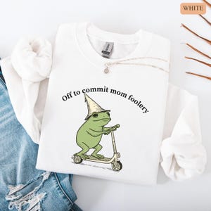 May include: White sweatshirt with the phrase "Off to commit mom foolery" and a cartoon frog wearing a wizard hat and riding a scooter. The frog is green, the hat is beige, and the scooter is beige and black.