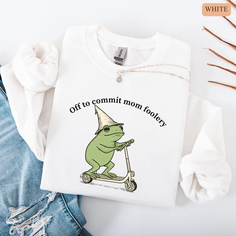 May include: White sweatshirt with the phrase "Off to commit mom foolery" and a cartoon frog wearing a wizard hat and riding a scooter. The frog is green, the hat is beige, and the scooter is beige and black.