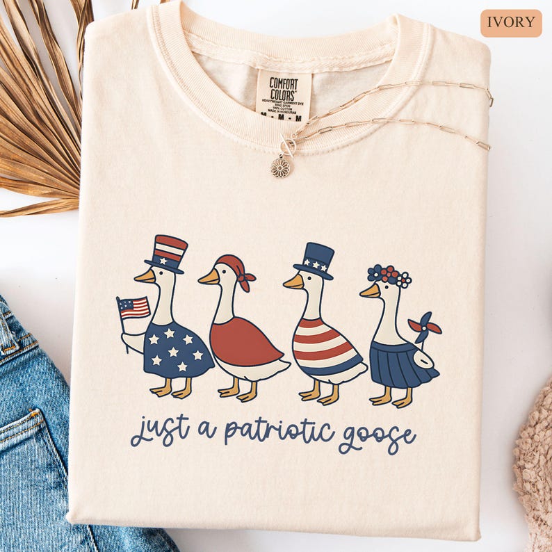 May include: Ivory-colored t-shirt featuring a patriotic goose design. Four geese are dressed in red, white, and blue outfits, with one holding an American flag. The text "just a patriotic goose" is written below the geese.