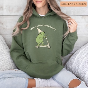 May include: Military green hoodie with a frog graphic riding a scooter and wearing a wizard hat and sunglasses. The text "Off to commit mom foolery" is printed above the frog. The hoodie has a drawstring hood and a front pocket.