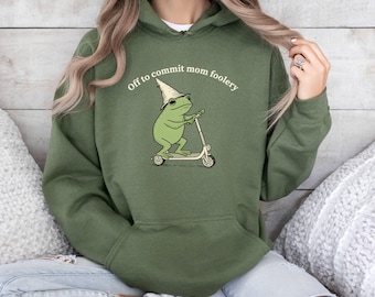 May include: Military green hoodie with a frog graphic riding a scooter and wearing a wizard hat and sunglasses. The text "Off to commit mom foolery" is printed above the frog. The hoodie has a drawstring hood and a front pocket.