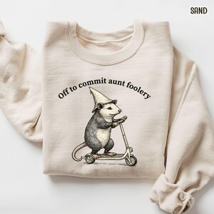 May include: A sand-colored sweatshirt with the phrase "Off to commit aunt foolery" printed in black. The graphic features an opossum wearing a wizard hat and riding a scooter. The sweatshirt is made of a soft material.