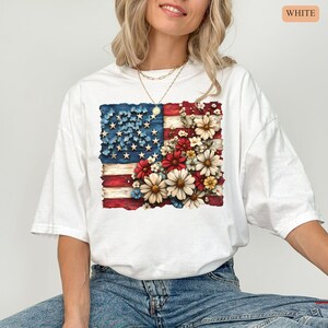 May include: White t-shirt featuring a distressed American flag design with floral accents. The flag's stars are blue, the stripes are red, and the flowers are red, white, and yellow. The word "WHITE" is printed in a small orange rectangle in the upper right corner.