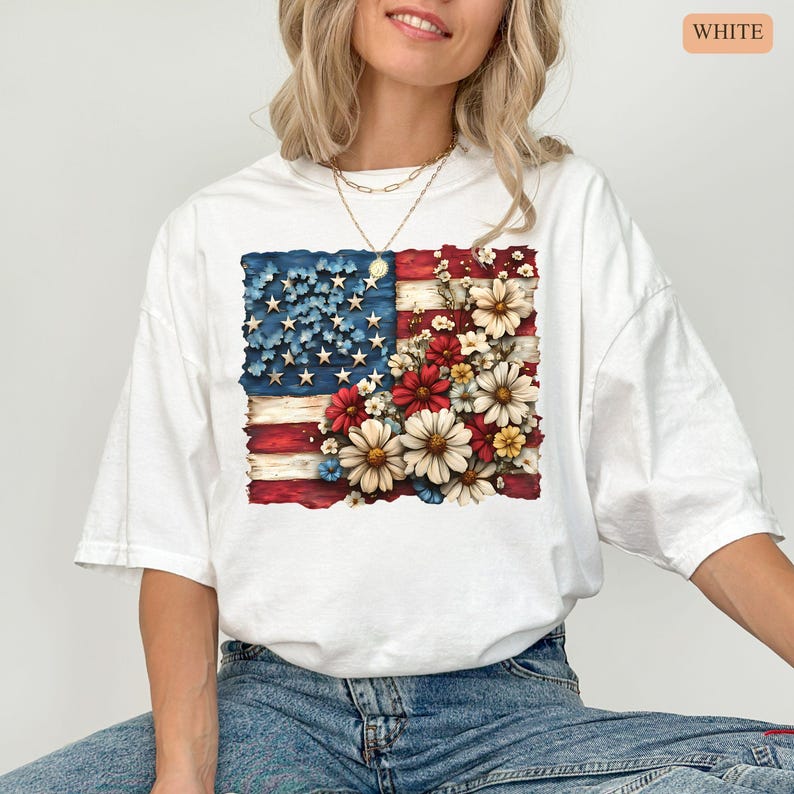 May include: White t-shirt featuring a distressed American flag design with floral accents. The flag's stars are blue, the stripes are red, and the flowers are red, white, and yellow. The word "WHITE" is printed in a small orange rectangle in the upper right corner.