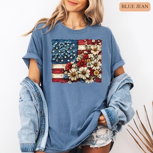 May include: Blue jean t-shirt featuring a distressed American flag design with a floral arrangement. The flag's stars are blue, and the stripes are red and white. The floral arrangement includes red, white, and yellow flowers.