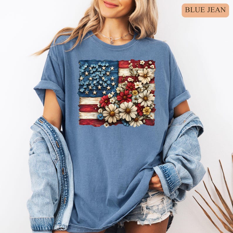 May include: Blue jean t-shirt featuring a distressed American flag design with a floral arrangement. The flag's stars are blue, and the stripes are red and white. The floral arrangement includes red, white, and yellow flowers.