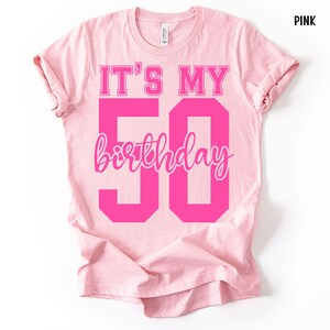 May include: A light pink short-sleeve t-shirt featuring the text IT'S MY 50 birthday in bold, dark pink block lettering and a cursive script font. The shirt has a crew neckline and a standard fit, with the word PINK printed in the top corner.