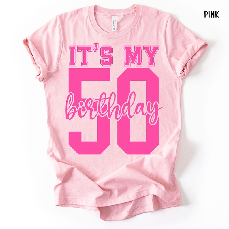 May include: A light pink short-sleeve t-shirt featuring the text IT'S MY 50 birthday in bold, dark pink block lettering and a cursive script font. The shirt has a crew neckline and a standard fit, with the word PINK printed in the top corner.