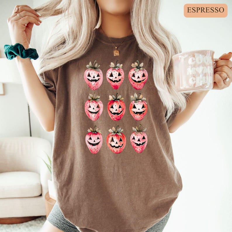 May include: Espresso-colored t-shirt featuring a repeating pattern of watercolor strawberry-jack-o'-lanterns. The strawberries have green tops and carved faces. The shirt is a relaxed fit and has short sleeves. The word "ESPRESSO" is printed in a rectangular box in the upper right corner.