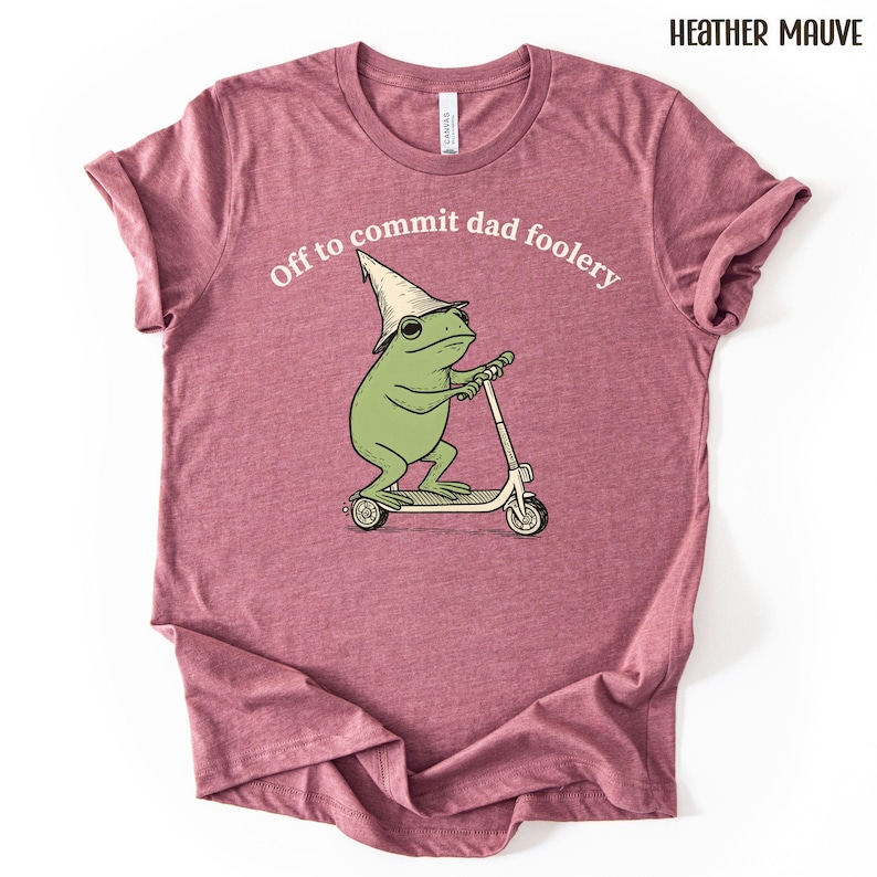 Puede incluir: Camiseta color malva jaspeado con un dibujo de una rana en un patinete, con un sombrero de mago. El texto "Off to commit dad foolery" est&aacute; sobre la rana. La camiseta es de material suave.