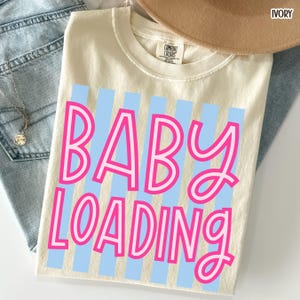 May include: Ivory-colored t-shirt with the text "BABY LOADING" in pink, outlined in white, over light blue vertical stripes. A tan hat and blue jeans are partially visible in the background. The shirt is made of a soft material.