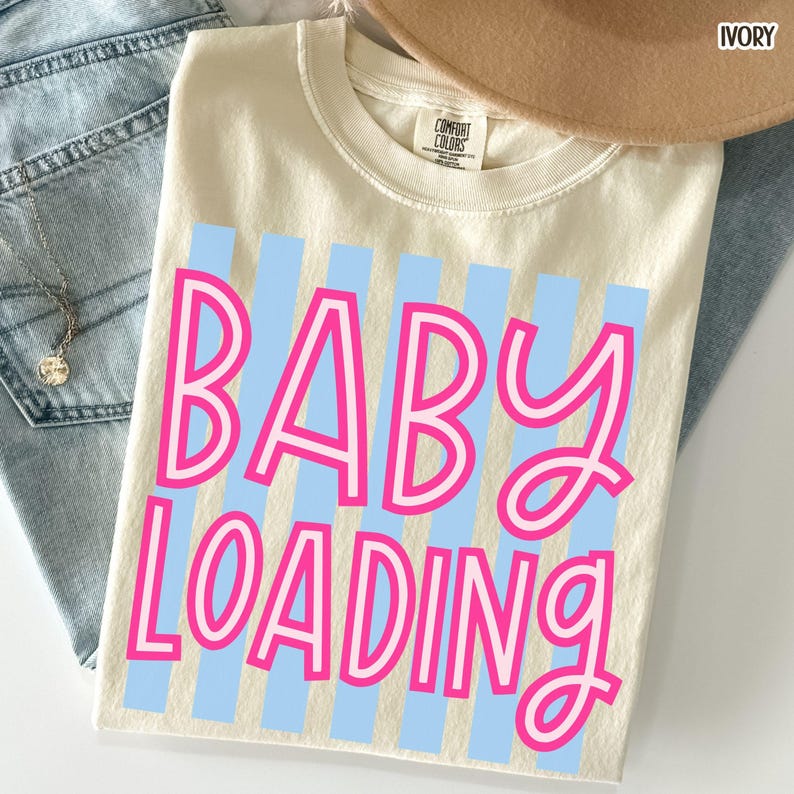 May include: Ivory-colored t-shirt with the text "BABY LOADING" in pink, outlined in white, over light blue vertical stripes. A tan hat and blue jeans are partially visible in the background. The shirt is made of a soft material.