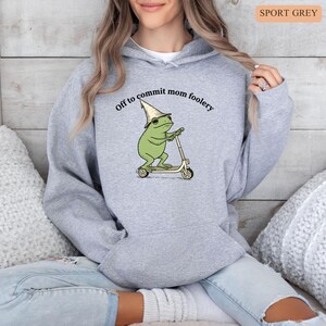 May include: Sport grey hoodie with a whimsical frog graphic riding a scooter and wearing a wizard hat. The text "Off to commit mom foolery" is arched above the frog. The hoodie is a light grey color.