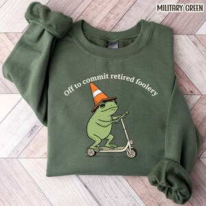 May include: A military green crewneck sweatshirt featuring a graphic of a green frog wearing an orange and white traffic cone as a hat while riding a kick scooter. The text above the frog reads, Off to commit retired foolery in white.