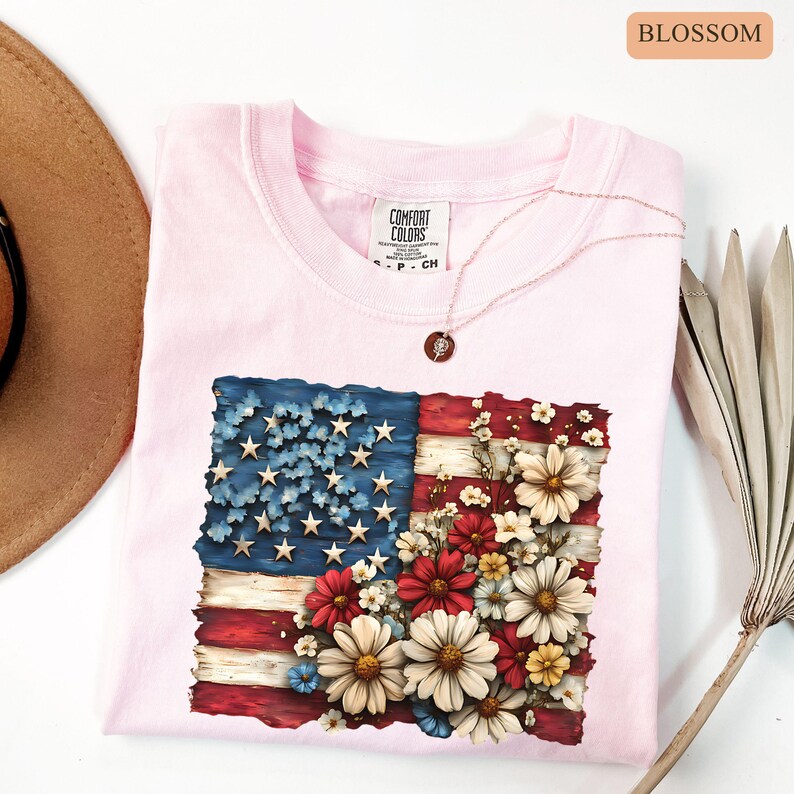 May include: A light pink t-shirt featuring a floral American flag design. The flag design incorporates red and white stripes, blue stars, and a bouquet of daisies. The shirt has a crew neck and short sleeves. A delicate necklace with a pendant rests on the shirt.