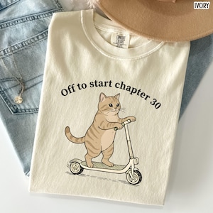 May include: Ivory-colored t-shirt with the text "Off to start chapter 30" above a cartoon cat riding a scooter. The cat is tan with brown stripes. The shirt is folded on top of blue jeans.