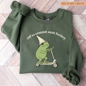 May include: Military green sweatshirt with the phrase "Off to commit aunt foolery." The graphic features a green frog wearing a wizard hat and riding a scooter. The sweatshirt is made of a soft material.
