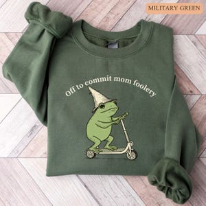 May include: Military green sweatshirt with the text "Off to commit mom foolery." The graphic features a green frog wearing a wizard hat and riding a scooter. The sweatshirt is a solid color.