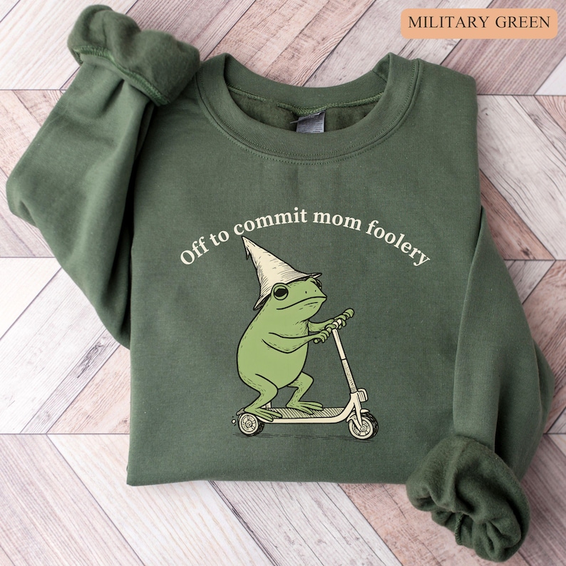 May include: Military green sweatshirt with the text "Off to commit mom foolery." The graphic features a green frog wearing a wizard hat and riding a scooter. The sweatshirt is a solid color with ribbed cuffs and a crew neck.