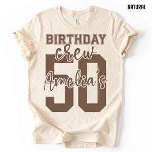 May include: A natural-colored short-sleeve t-shirt featuring dark brown text that reads BIRTHDAY crew Amelia's 50. The design uses a mix of block and script fonts, with the number 50 prominently displayed in a large, bold, dark brown block font.