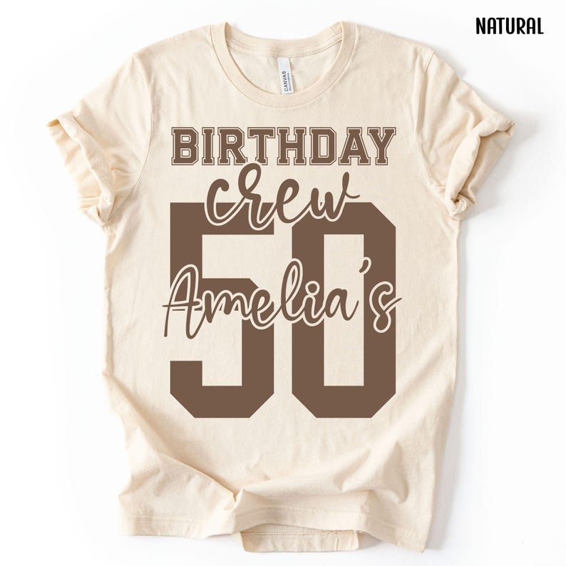 May include: A natural-colored short-sleeve t-shirt featuring dark brown text that reads BIRTHDAY crew Amelia's 50. The design uses a mix of block and script fonts, with the number 50 prominently displayed in a large, bold, dark brown block font.