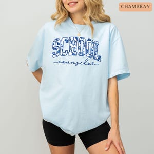 May include: Light blue t-shirt with the words "SCHOOL counselor" in navy blue. The word "SCHOOL" is filled with a floral pattern. The shirt is a crew neck and has short sleeves. The model is wearing black shorts.
