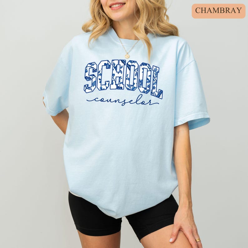 May include: Light blue t-shirt with the words "SCHOOL counselor" in navy blue. The word "SCHOOL" is filled with a floral pattern. The shirt is a crew neck and has short sleeves. The model is wearing black shorts.