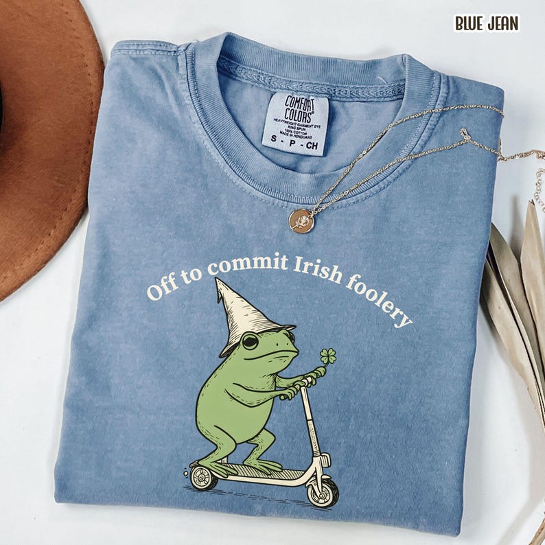 May include: Blue jean Comfort Colors t-shirt with the phrase "Off to commit Irish foolery" and a cartoon frog riding a scooter. The frog wears a pointed hat and holds a four-leaf clover. A gold necklace is draped over the shirt.