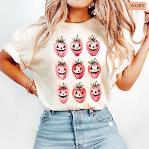 May include: Ivory-colored t-shirt featuring a repeating pattern of pink strawberries with carved jack-o'-lantern faces. The strawberries have green stems and leaves. The shirt is a light, neutral color, suitable for Halloween.