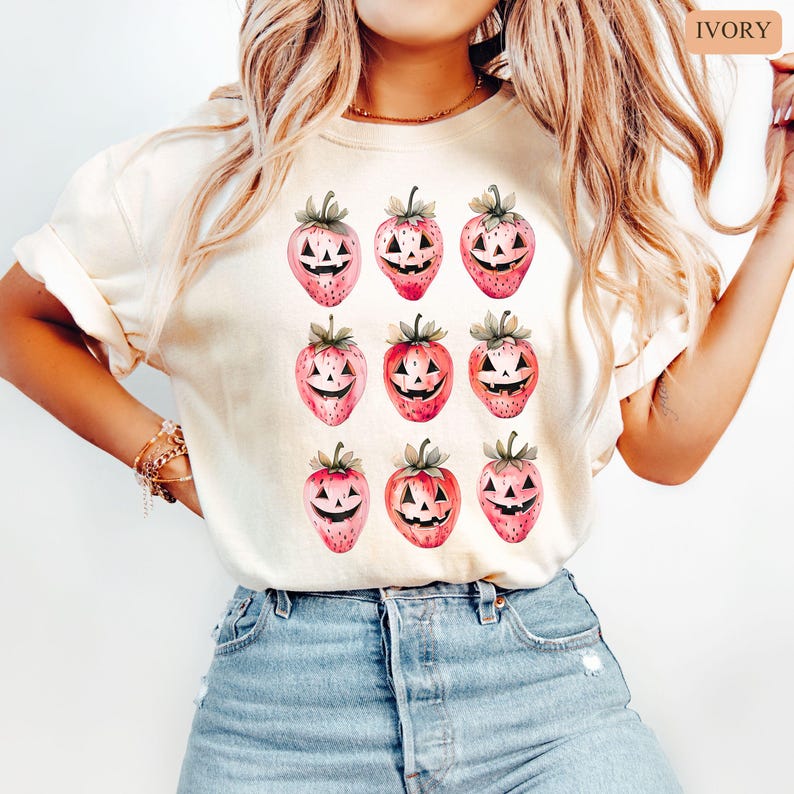 May include: Ivory-colored t-shirt featuring a repeating pattern of pink strawberries with carved jack-o'-lantern faces. The strawberries have green stems and leaves. The shirt is a light, neutral color, suitable for Halloween.