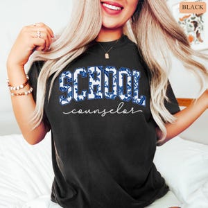May include: Black t-shirt with the words "SCHOOL counselor" in a blue and white floral design. The word "SCHOOL" is in a bold, block letter style, while "counselor" is in a cursive font.
