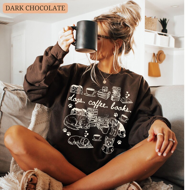 May include: Dark chocolate-colored sweatshirt with the text "dogs. coffee. books." and illustrations of dogs, books, and coffee cups. A black mug is held up to the face. The sweatshirt is a comfortable, casual style.
