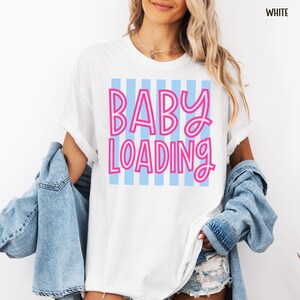 May include: White t-shirt with the text "BABY LOADING" in pink, outlined in white, over a light blue striped background. The shirt is a casual style, with rolled-up sleeves. The word "WHITE" is in the top right corner.