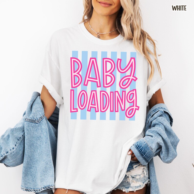 May include: White t-shirt with the text "BABY LOADING" in pink, outlined in white, over a light blue striped background. The shirt is a casual style, with rolled-up sleeves. The word "WHITE" is in the top right corner.