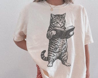 May include: Ivory-coloured t-shirt with a black and white illustration of a cat reading a book. The cat is standing upright, holding the book open. The shirt has short sleeves and a crew neck. The word "IVORY" is printed at the top.