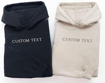 May include: Two long-sleeve hooded sweatshirts, one in black and one in beige, each featuring the words CUSTOM TEXT printed in a simple serif font across the chest area. Both garments include a hood with matching drawstrings and ribbed cuffs.