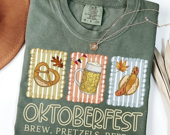 May include: A moss green t-shirt with an Oktoberfest design. The design includes a pretzel, a beer stein, and a hot dog, with the words "OKTOBERFEST BREW, PRETZELS, REPEAT". The shirt has a crew neck and short sleeves.