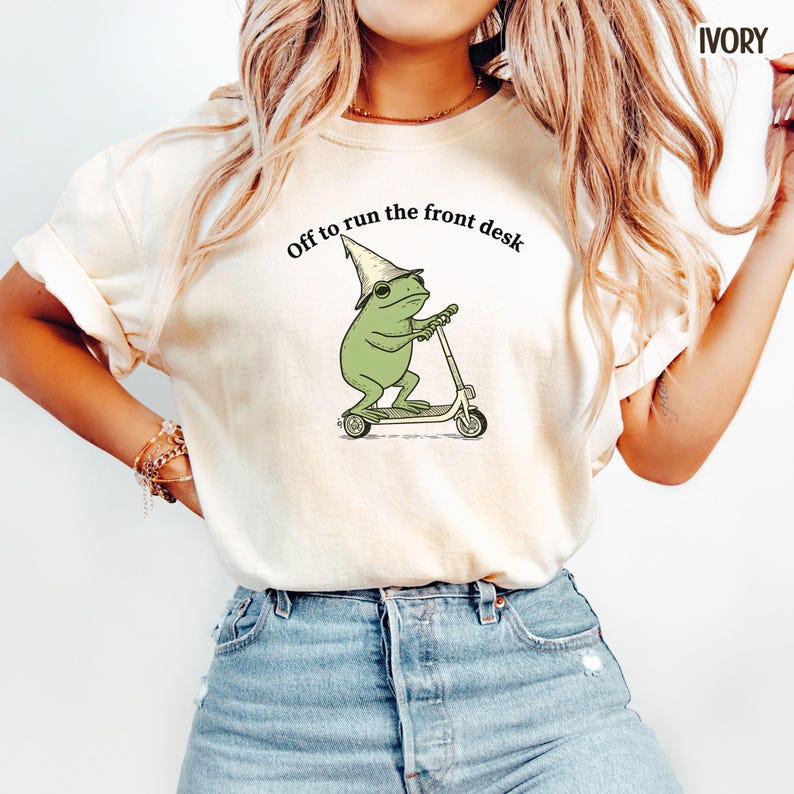 K&ouml;nnte beinhalten: Elfenbeinfarbenes T-Shirt mit einer Illustration eines gr&uuml;nen Frosches auf einem Roller, der einen Zaubererhut tr&auml;gt. Der Text "Off to run the front desk" ist dar&uuml;ber gedruckt. Das Shirt ist aus weichem Material.