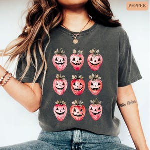 May include: Dark gray t-shirt featuring a repeating pattern of strawberries with jack-o'-lantern faces. The strawberries are pink and red with green stems and leaves. The shirt is a comfortable, casual style.