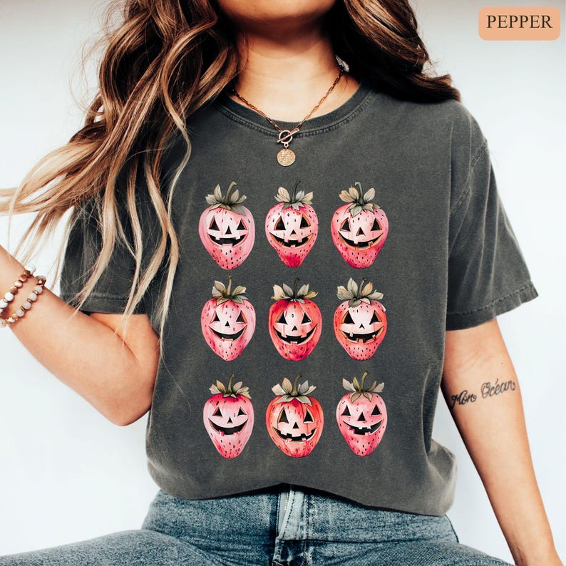 May include: Dark gray t-shirt featuring a repeating pattern of strawberries with jack-o'-lantern faces. The strawberries are pink and red with green stems and leaves. The shirt is a comfortable, casual style.
